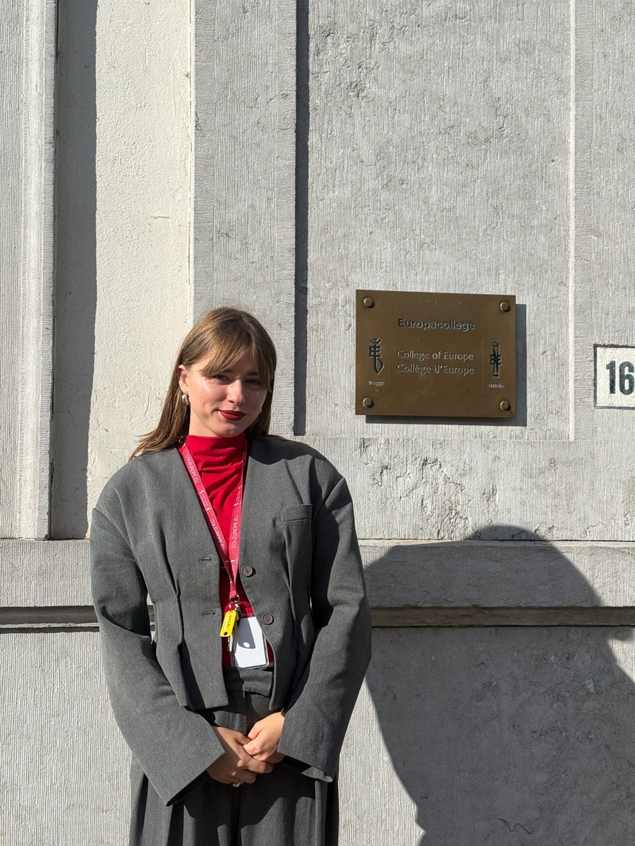 Taya, in a grey suit and red top, standing beside the College of Europe&rsquo;s brass nameplate